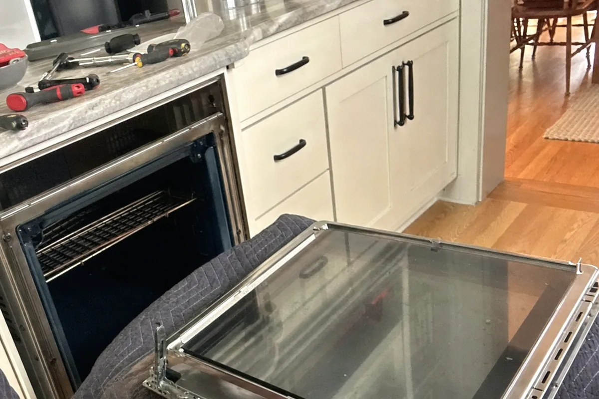 Inner door glass held out of a built-in oven, the hinge frame and rack rails visible in the opening behind it