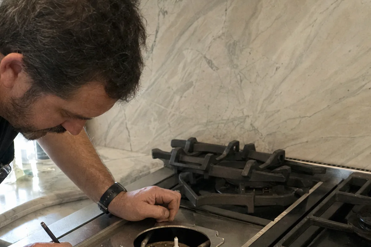 A wide stainless range set into a stone counter, seen from the side while one of its burners is worked on